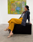 Woman sitting on a black ottoman wearing mustard yellow pants in an art gallery with a colorful abstract painting in the background.