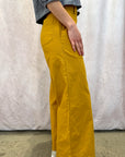 Woman wearing mustard yellow pants and a gray shirt against a white curtain background